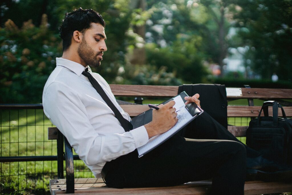 A thoughtful businessman writing notes on a clipboard while sitting on a park bench.